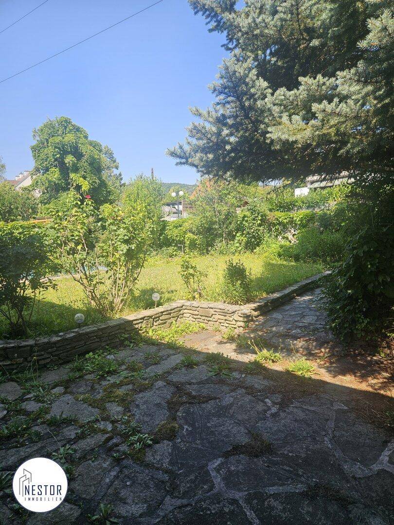 Verwilderter Gartenbereich mit Steinplattenweg und niedrigen Steinmauern unter strahlendem Sonnenschein.