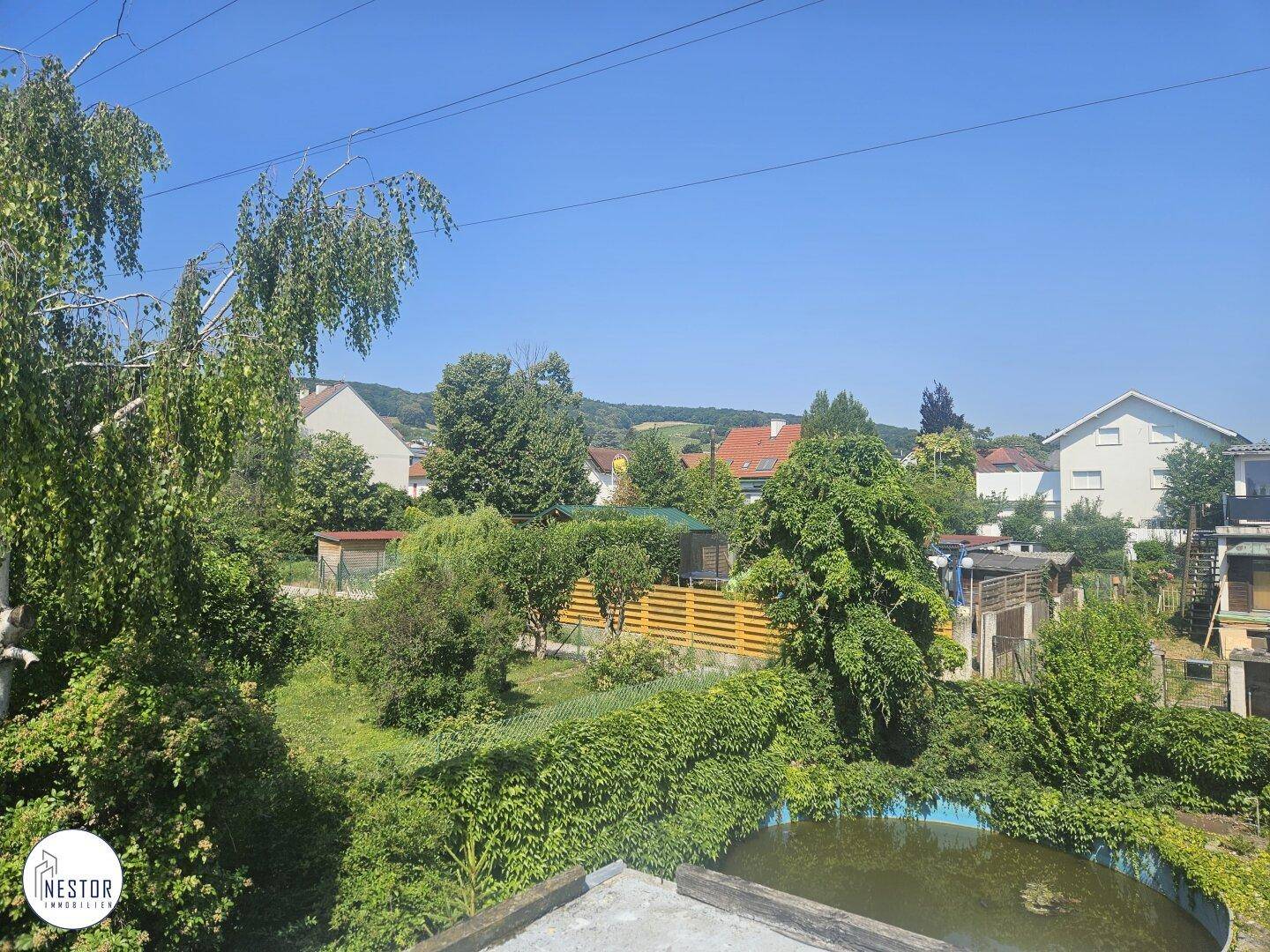 Weitblick über einen verwilderten Garten mit Pool und die umliegende Landschaft mit Häusern.
