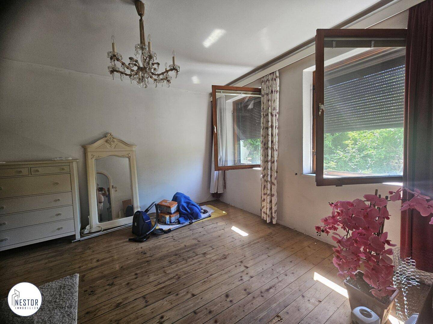 Helles Schlafzimmer mit Holzdielenboden, Kronleuchter und einem großen Spiegel, mit Blick ins Grüne.