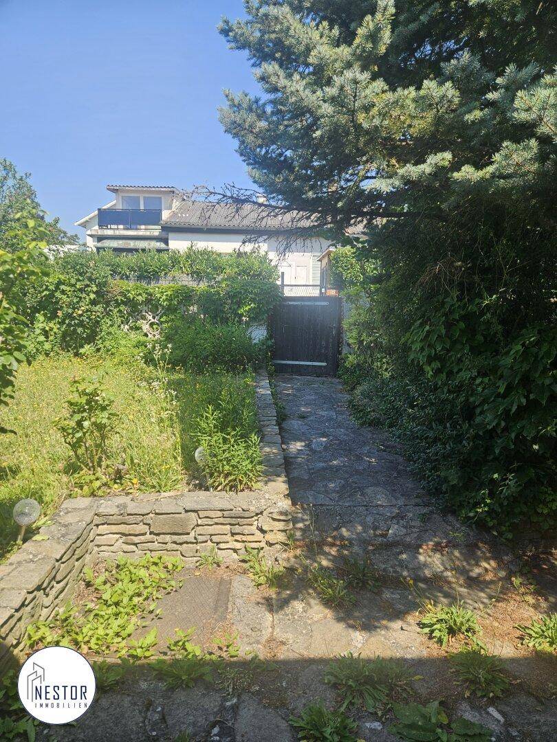 Steinweg führt durch einen verwilderten Garten zu einem Haus mit Balkon im Hintergrund.