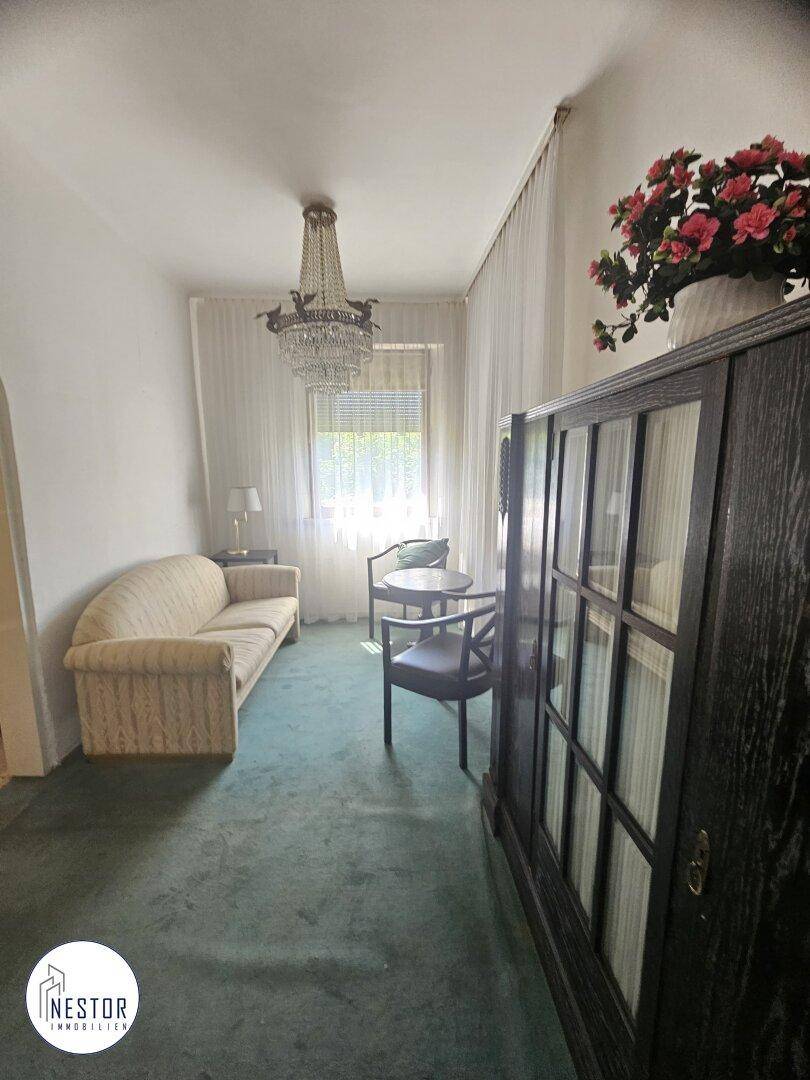 Gemütliches Wohnzimmer mit Teppichboden, Sofa und Kronleuchter, hell durch das Fenster.