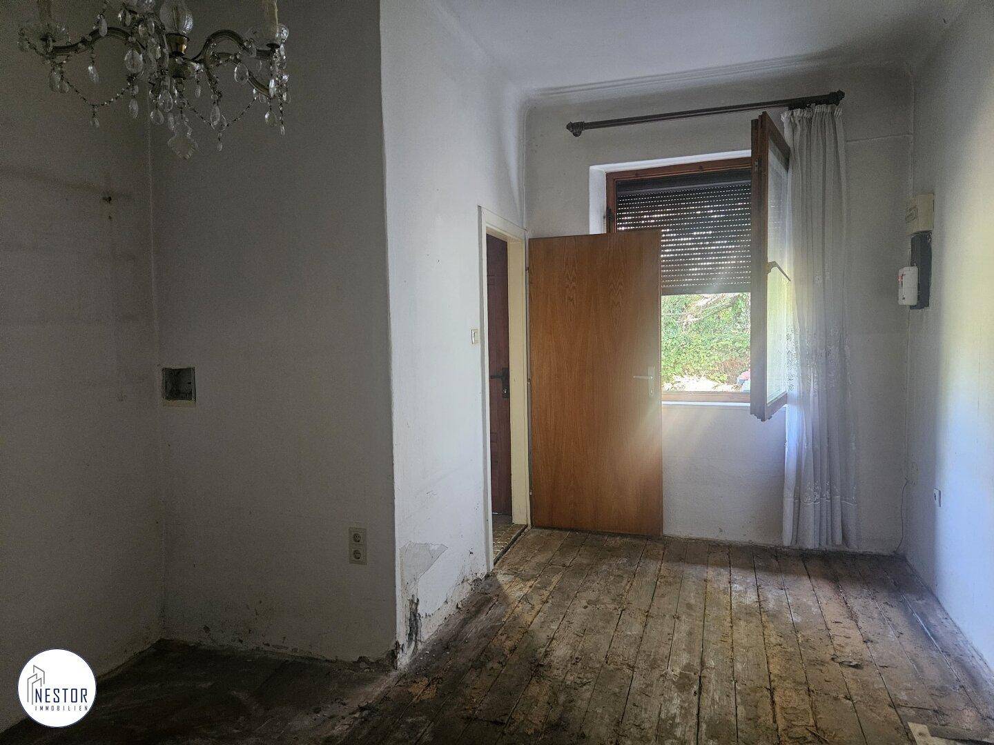 Raum mit Holzdielenboden und Fenster, das auf den Garten blickt, in renovierungsbedürftigem Zustand.