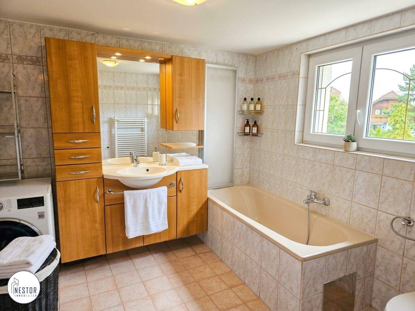 Helles Badezimmer mit Badewanne, Holzmöbeln und Fenster mit Blick ins Grüne.