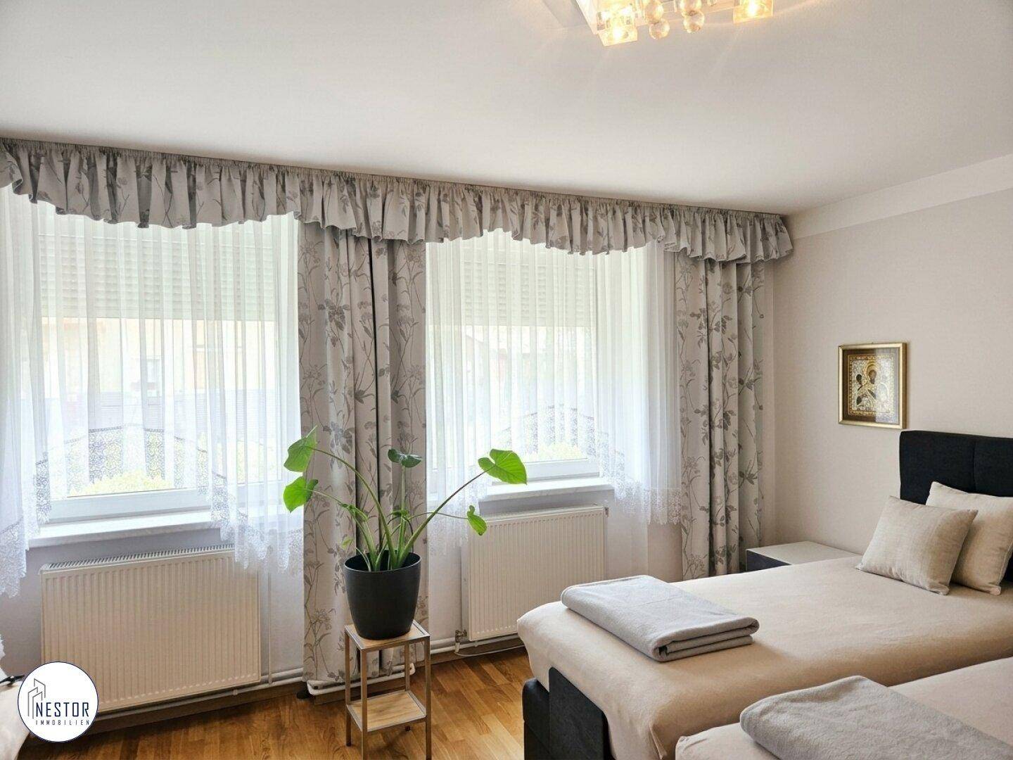 Helles Schlafzimmer mit zwei Einzelbetten, großen Fenstern und floral gemusterten Vorhängen.