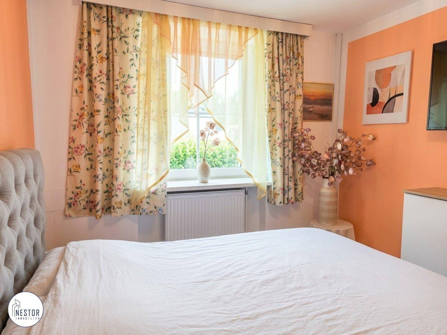 Schlafzimmer mit floralen Vorhängen, orangefarbener Akzentwand und Blick ins Grüne.