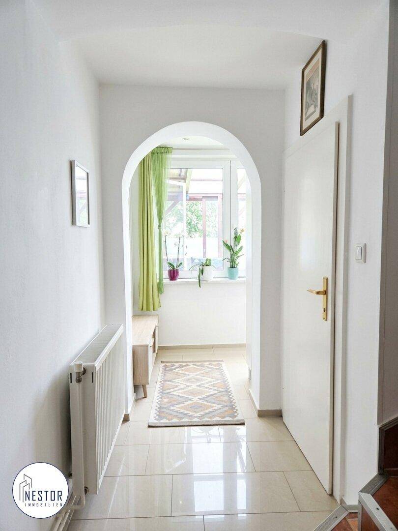Heller Flur mit Fliesenboden, weiß gestrichenen Wänden und einem Bogen zum Fensterbereich.
