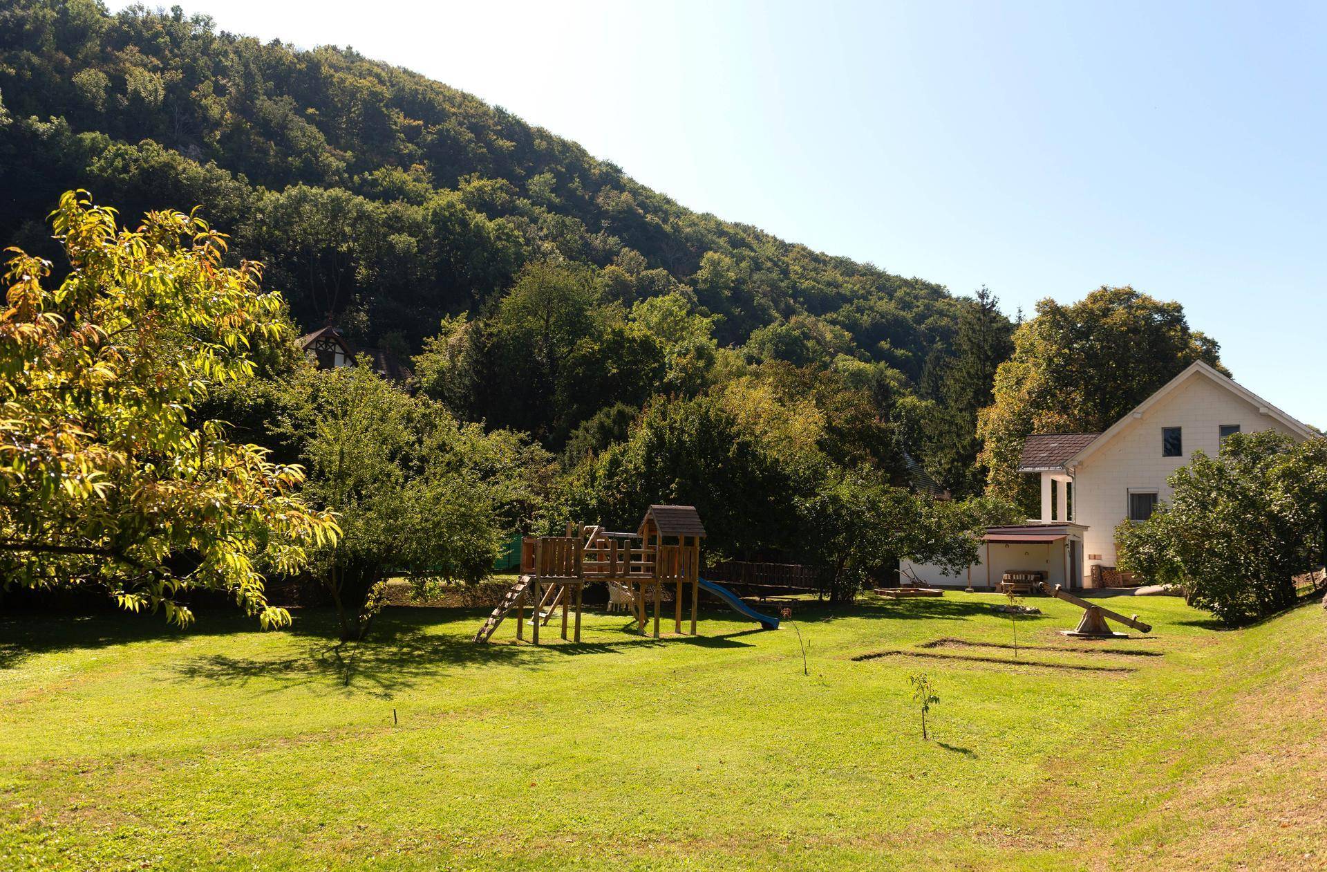 Weitläufiger Garten mit Spielplatz und Rutsche, umgeben von einem bewaldeten Hügel und einem Haus.