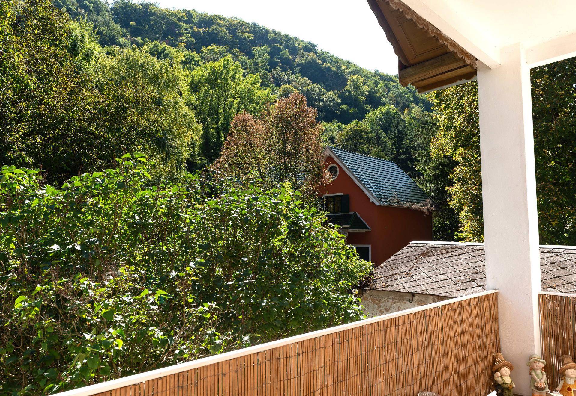 Balkon mit Blick auf eine bewaldete Hügellandschaft und ein rotes Haus im Hintergrund.