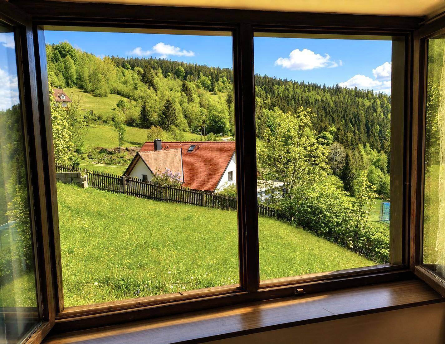 Fensterblick auf die grüne Hügellandschaft mit einem weiteren Haus und Wald im Hintergrund.