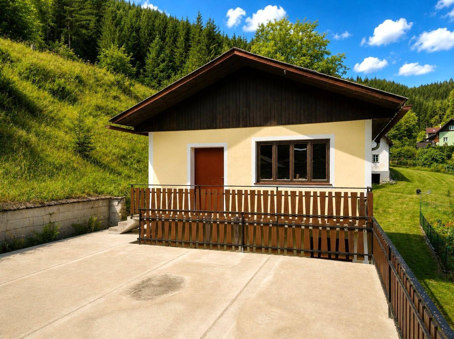 Kleines Gebäude mit Beton-Terrasse und Holzzaun, umgeben von grüner Natur.