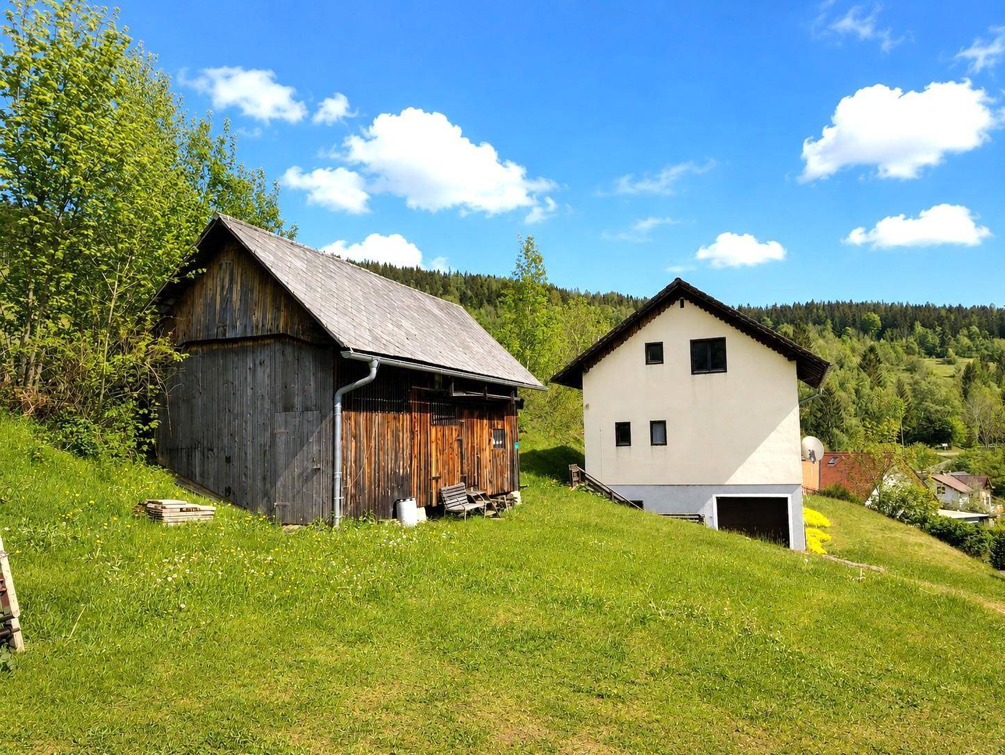 Ländliches Anwesen mit Haupthaus und rustikaler Holzscheune auf einem grünen Hang.