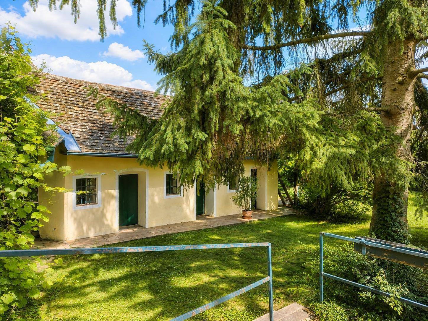 Charmantes gelbes Haus mit grünen Türen, umgeben von einem gepflegten Garten und hohen Bäumen.
