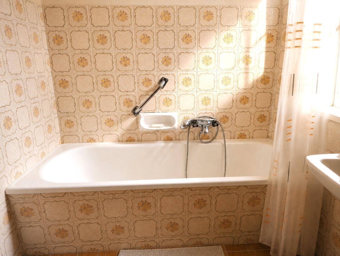 Badezimmer mit Badewanne, Haltegriff und gemusterten Fliesen im Retro-Stil.
