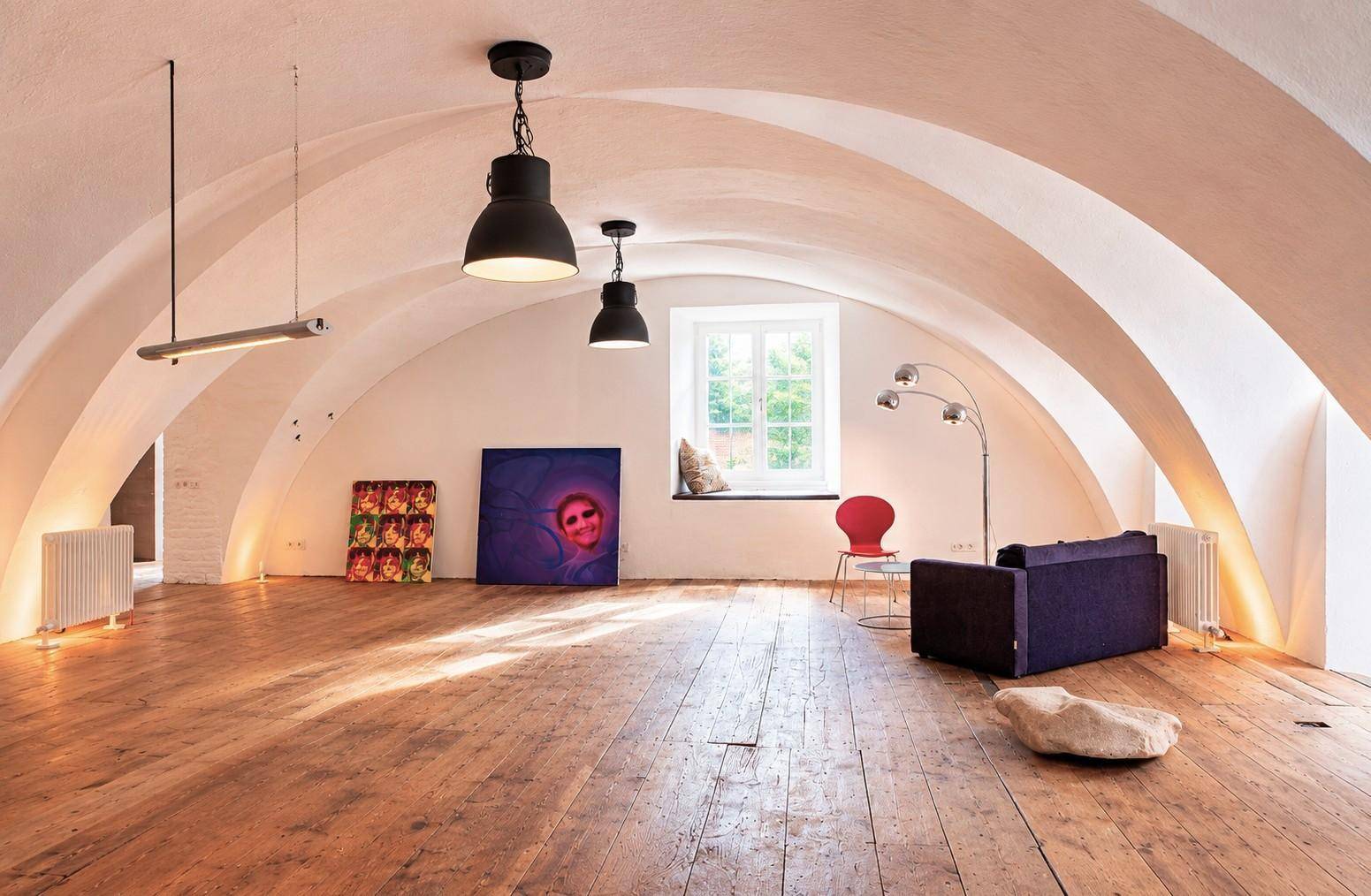 Heller Wohnraum mit markanten Gewölbedecken, breiten Holzdielen und einem großen Fenster, dekoriert mit Kunstwerken.