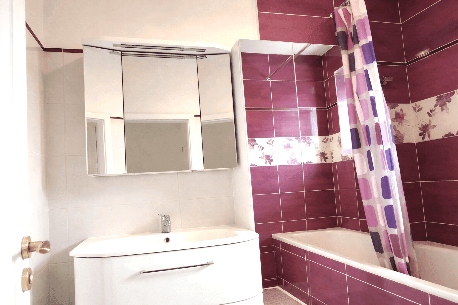 Badezimmer mit lila und weißen Fliesen, Badewanne mit Duschvorhang und Spiegelschrank.