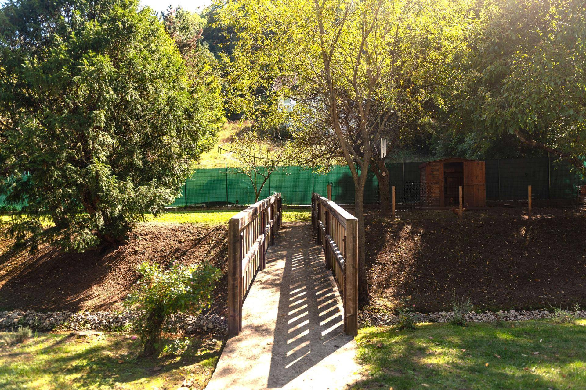 Holzbrücke über einen kleinen Bach in einer sonnigen, grünen Landschaft mit Bäumen.