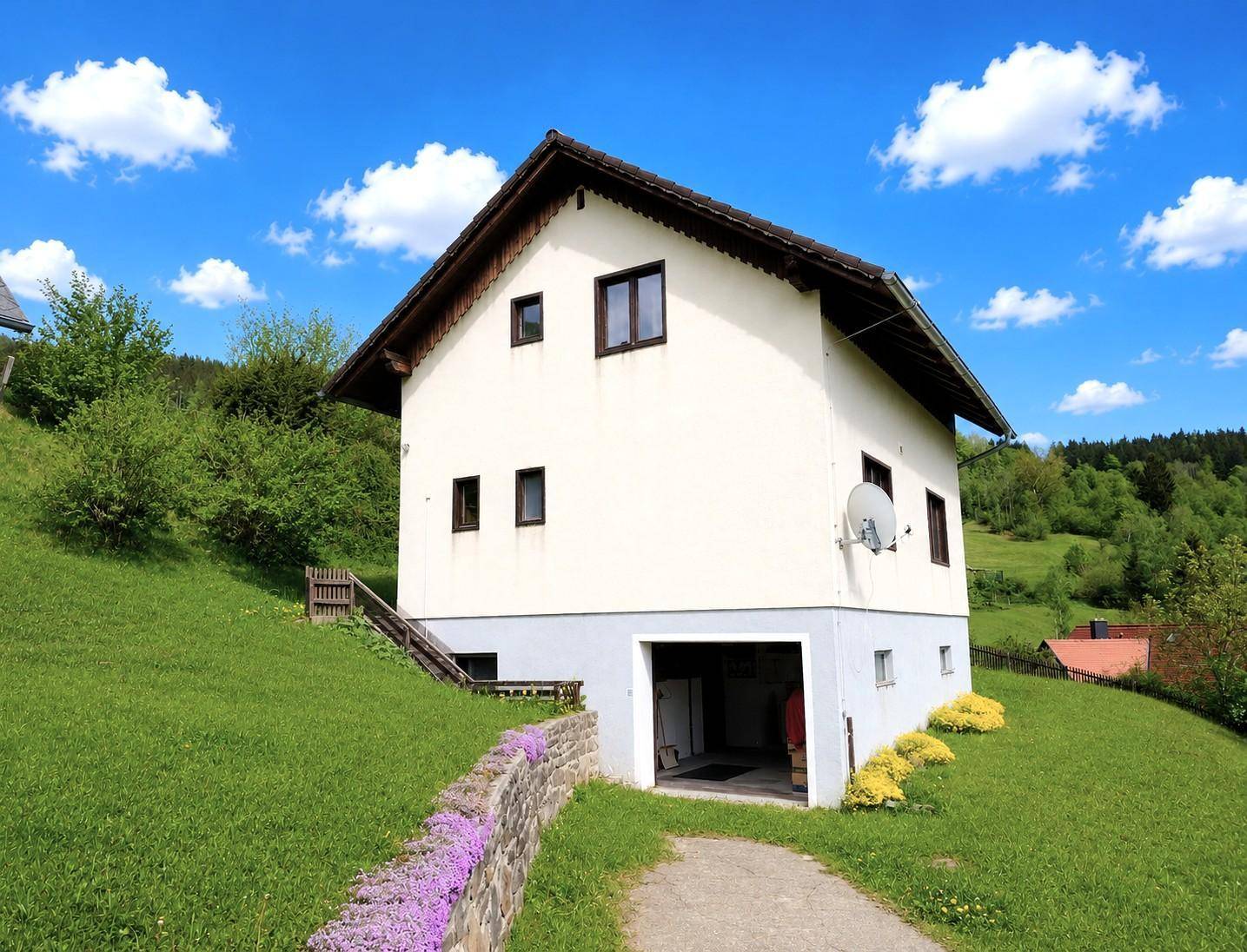 Weißes Einfamilienhaus mit integrierter Garage und blühenden Pflanzen am Hang.