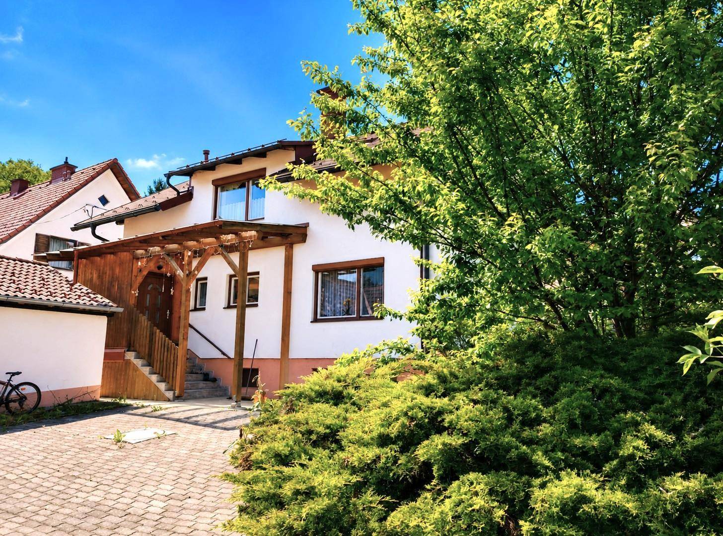 Eingangsbereich des Hauses mit Holzpergola und gepflasterter Zufahrt, umgeben von Grün.