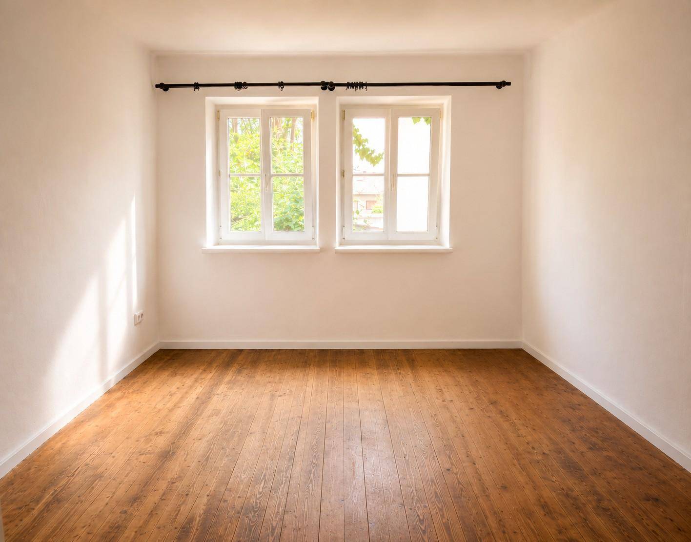 Helles, leeres Zimmer mit Holzdielenboden und zwei Fenstern, die viel Tageslicht hereinlassen.