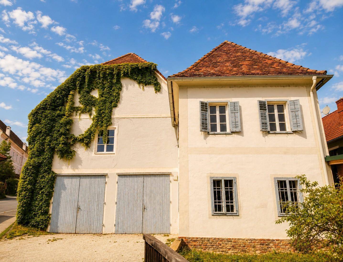 Vorderansicht eines historischen Gebäudes mit blauen Garagentoren, Fensterläden und einem roten Ziegeldach.