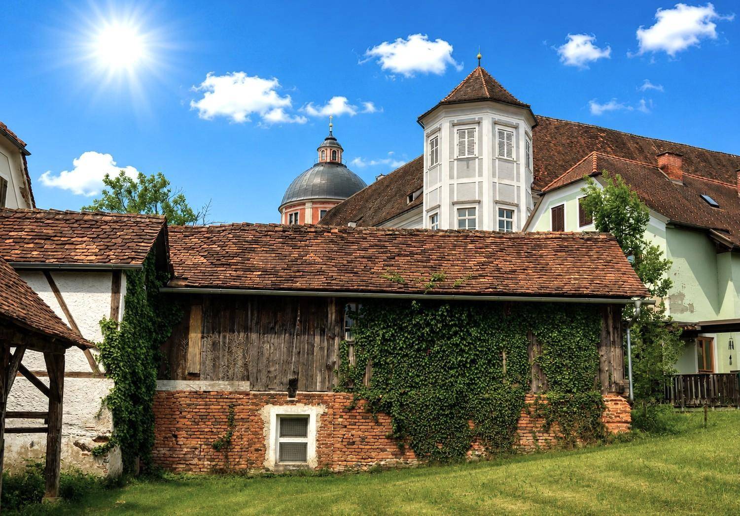 Sonnige Ansicht alter Gebäude mit roten Ziegeldächern und grünen Wiesen unter einem strahlend blauen Himmel.