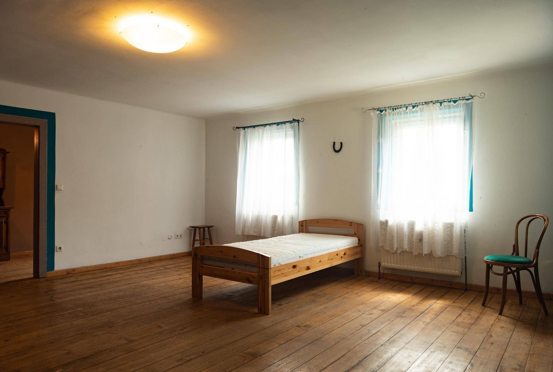 Helles Zimmer mit Holzdielenboden, einem Bett und zwei Fenstern, durchflutet von natürlichem Licht.