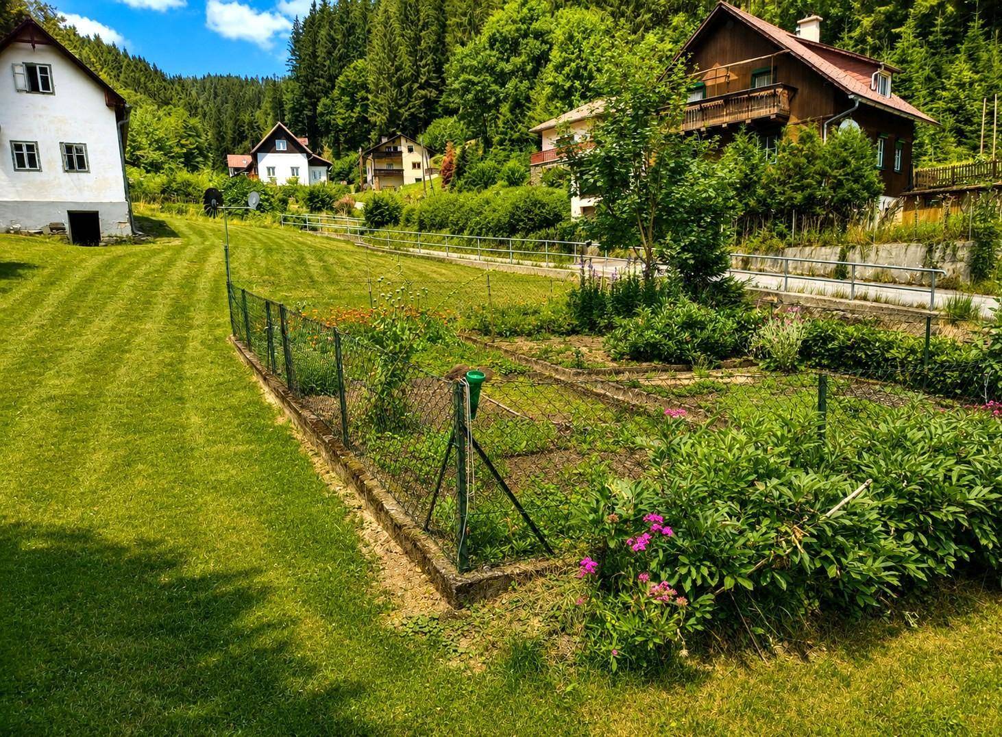Eingezäunter Gartenbereich mit Blumenbeeten und Blick auf weitere Häuser in der Ferne.