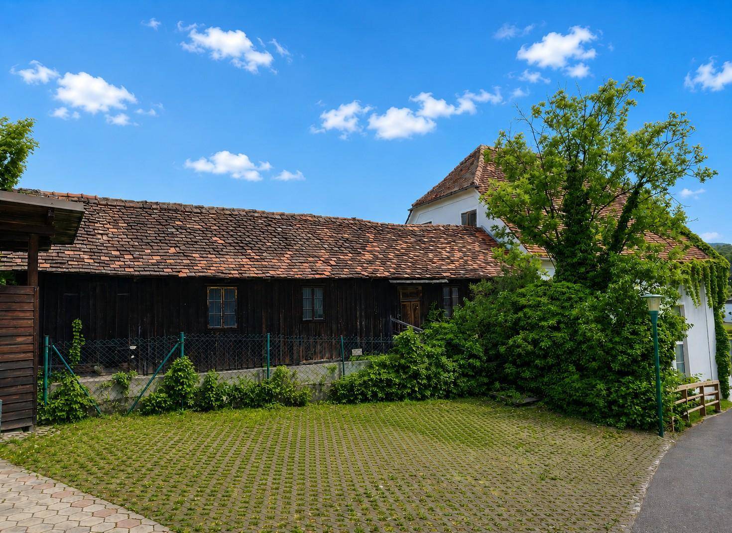 Ländliche Ansicht eines Holzgebäudes mit Ziegeldach, umgeben von Grünflächen und einem gepflasterten Bereich.
