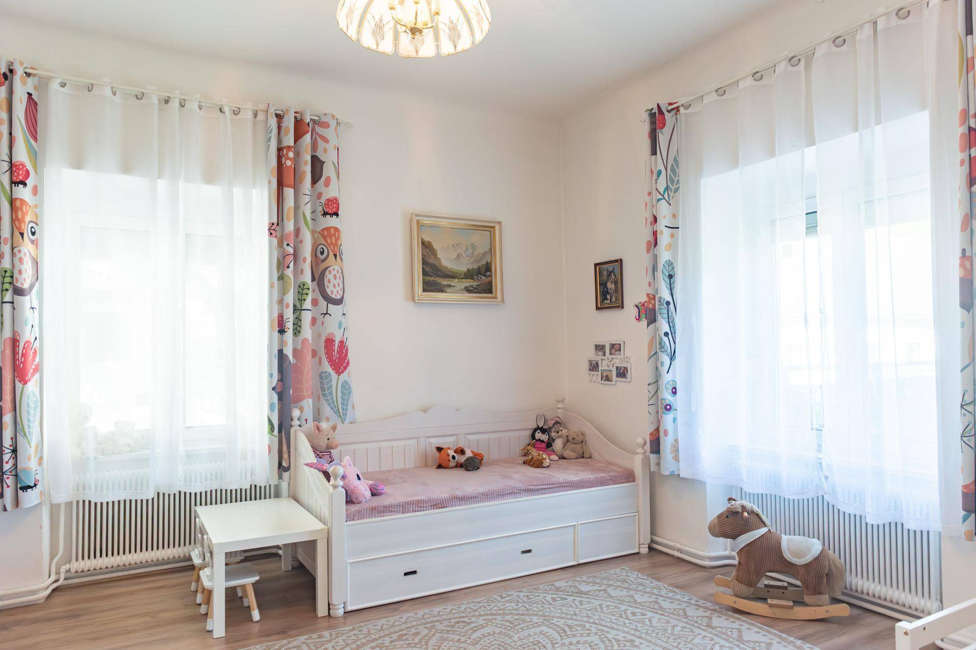 Kinderzimmer mit hellem Holzboden, zwei Fenstern und einem Bett, dekoriert mit bunten Vorhängen.