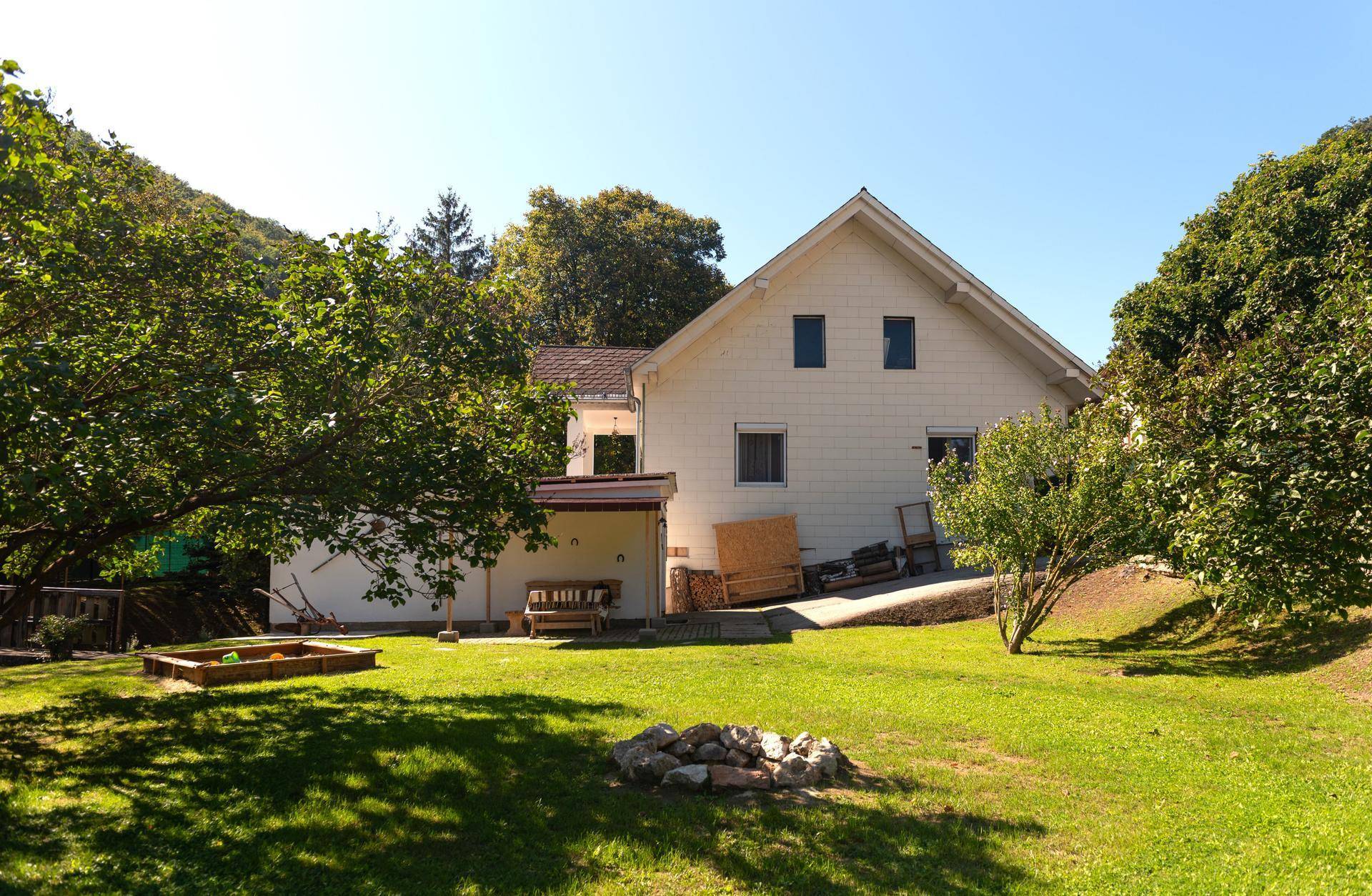 Rückansicht eines Hauses mit Garten, Feuerstelle und Holzschuppen, umgeben von viel Grün.
