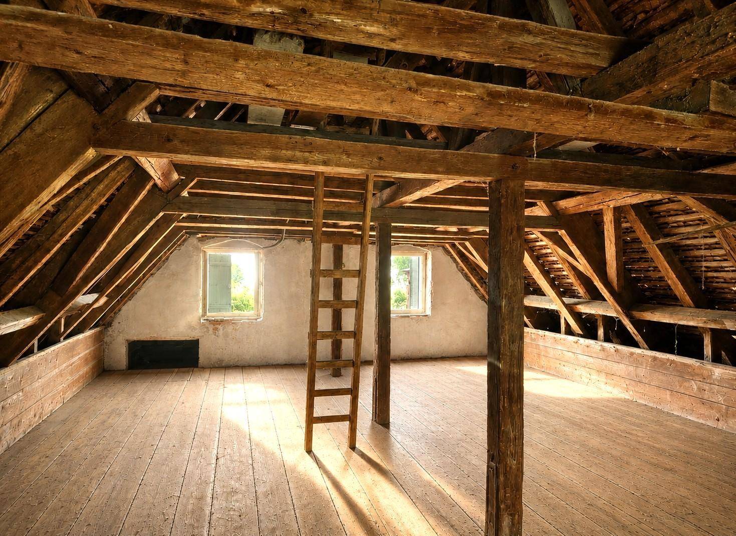 Dachboden mit sichtbaren Holzbalken, einer Holzleiter und zwei Fenstern, die den Raum erhellen.