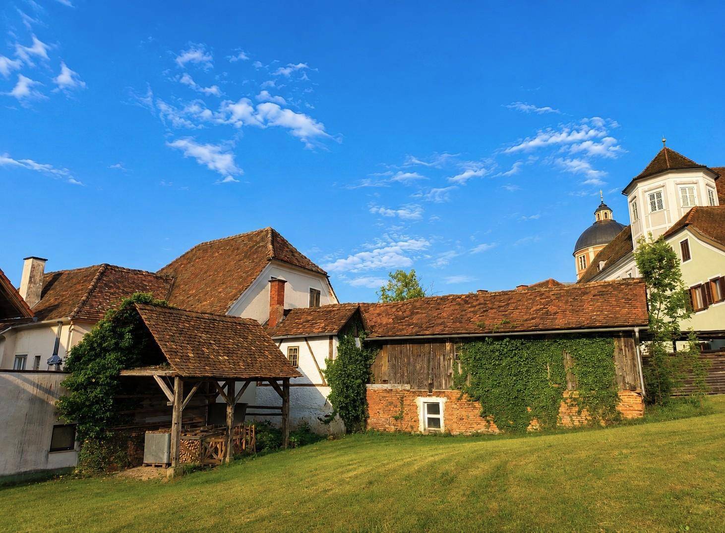 Malerische Ansicht alter Gebäude mit roten Ziegeldächern und grünen Wiesen unter einem strahlend blauen Himmel.