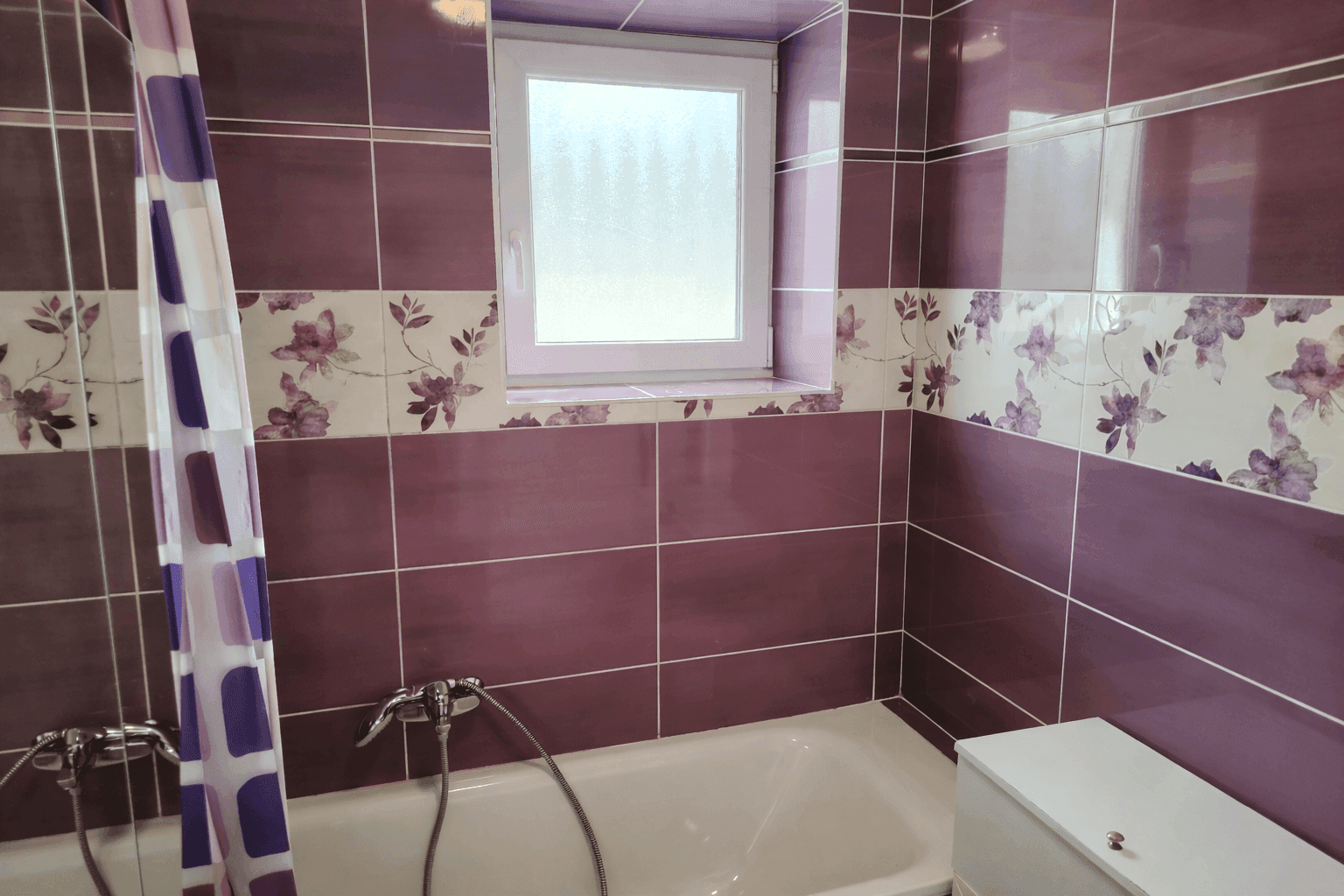 Badezimmer mit lila und weißen Fliesen, Badewanne und einem Fenster für Tageslicht.