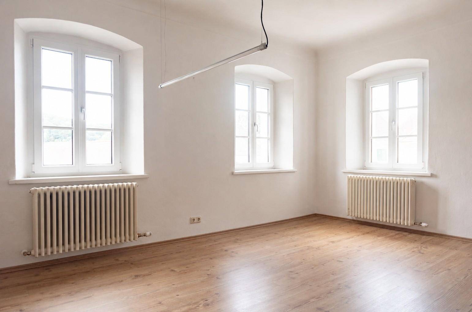 Heller, leerer Raum mit weißen Wänden, hellem Laminatboden und drei großen Fenstern mit Heizkörpern darunter.