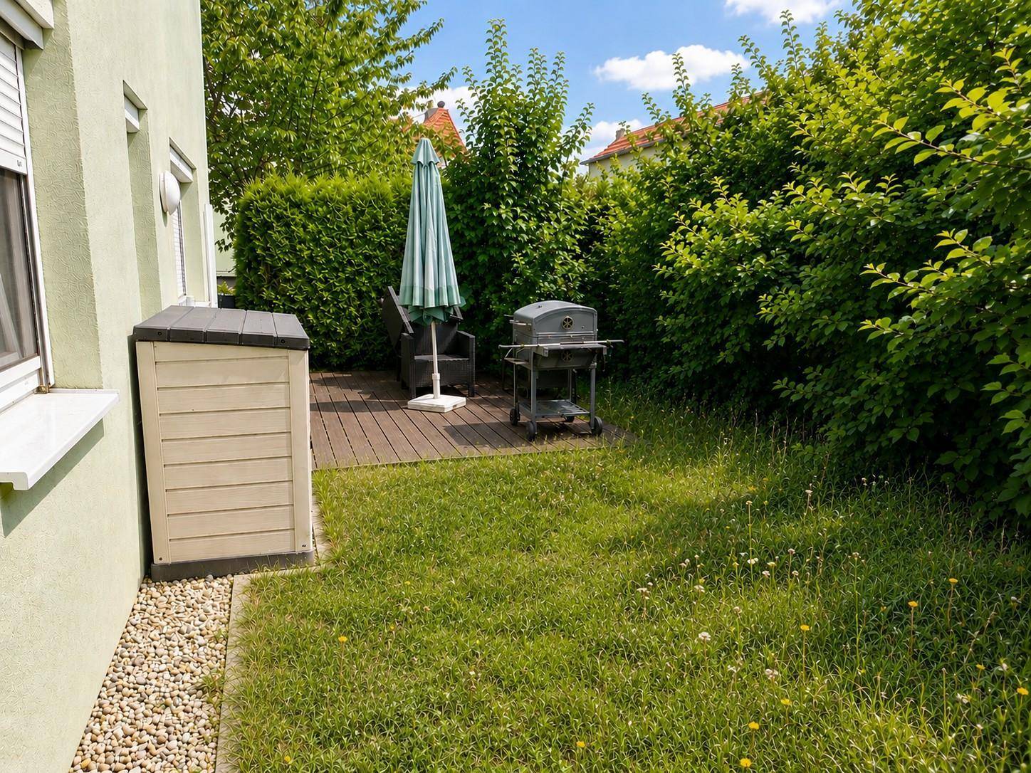 Sonniger Gartenbereich mit Rasen, Grill und kleiner Holzterrasse.