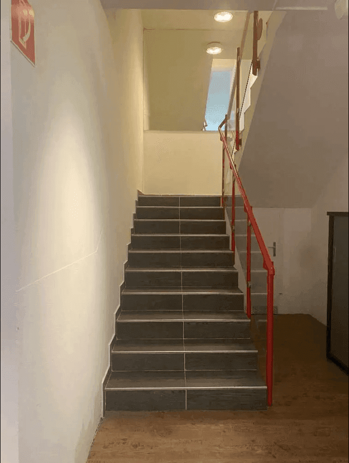 Treppenhaus mit dunklen Fliesenstufen und einem auffälligen roten Geländer, das nach oben führt.