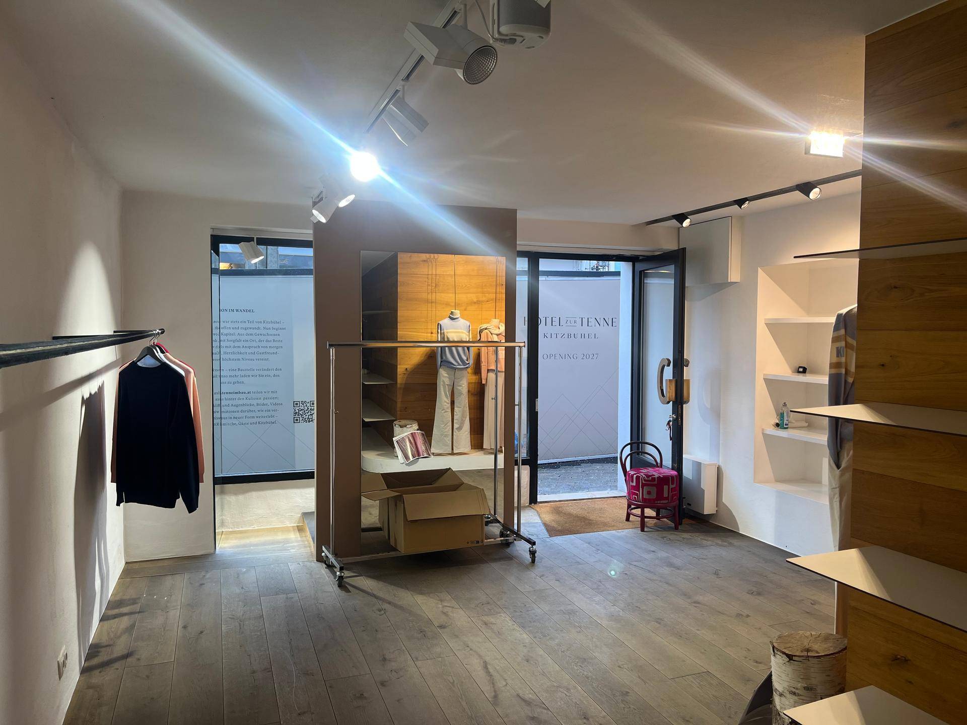 Heller Ladenbereich mit Holzboden, Regalen und Kleiderständern, durch die Eingangstür ist die Straße sichtbar.