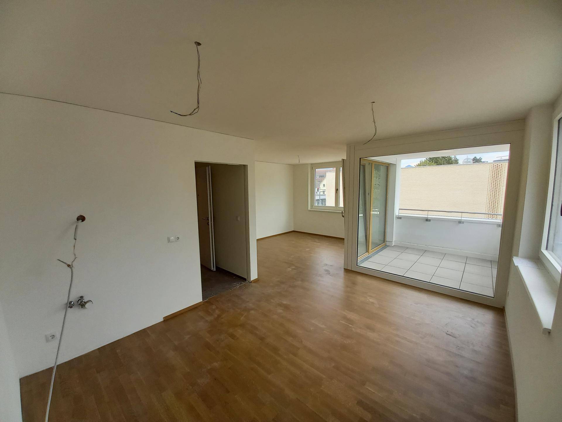 Geräumiges, leeres Wohnzimmer mit Holzboden und Zugang zu einem gefliesten Balkon, der viel Tageslicht hereinlässt.