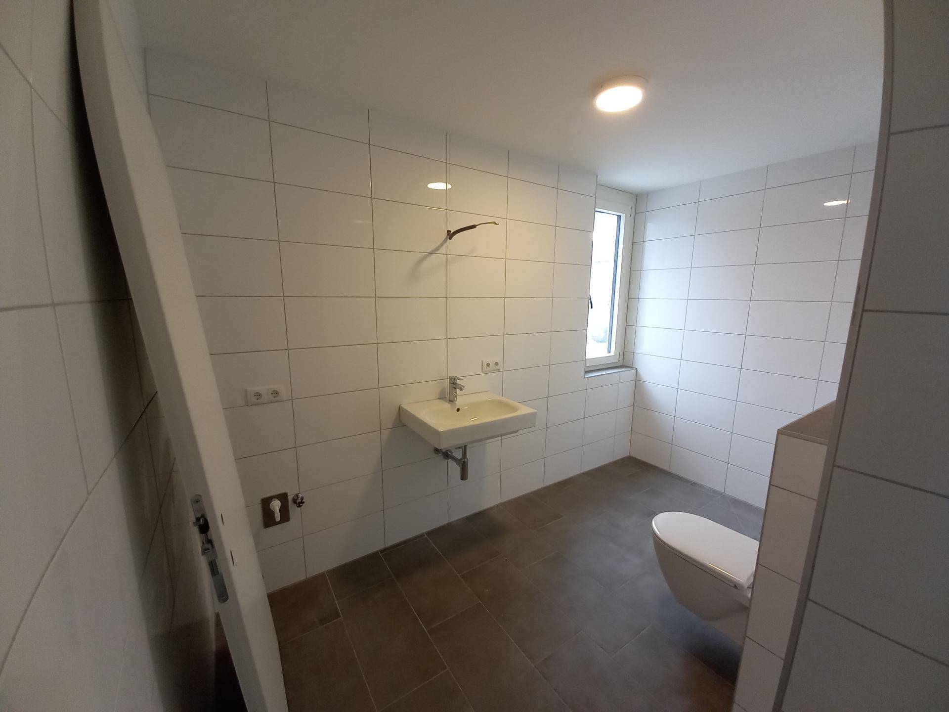 Modernes Badezimmer mit weißen Wandfliesen, grauem Boden und wandhängendem WC sowie einem Fenster für Tageslicht.