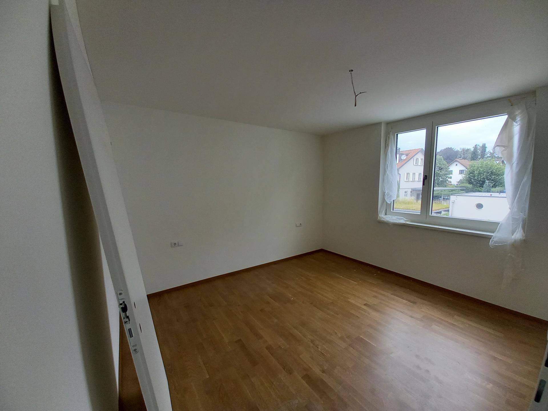 Leeres Schlafzimmer mit hellem Holzboden und einem großen Fenster, das viel natürliches Licht in den Raum bringt.