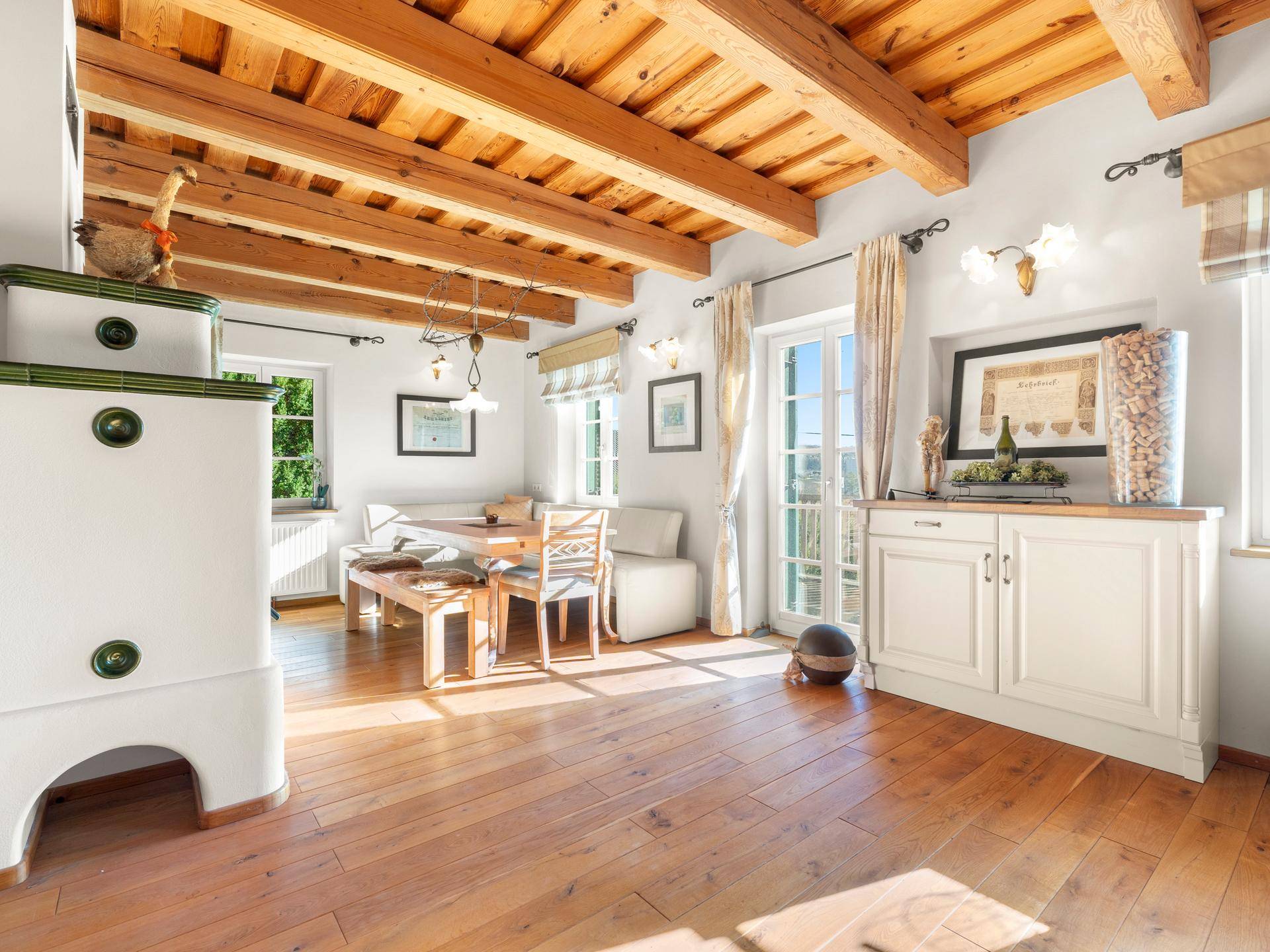 Rustikaler Essbereich mit Kachelofen, Holzbalken und Zugang zu einem sonnigen Außenbereich.