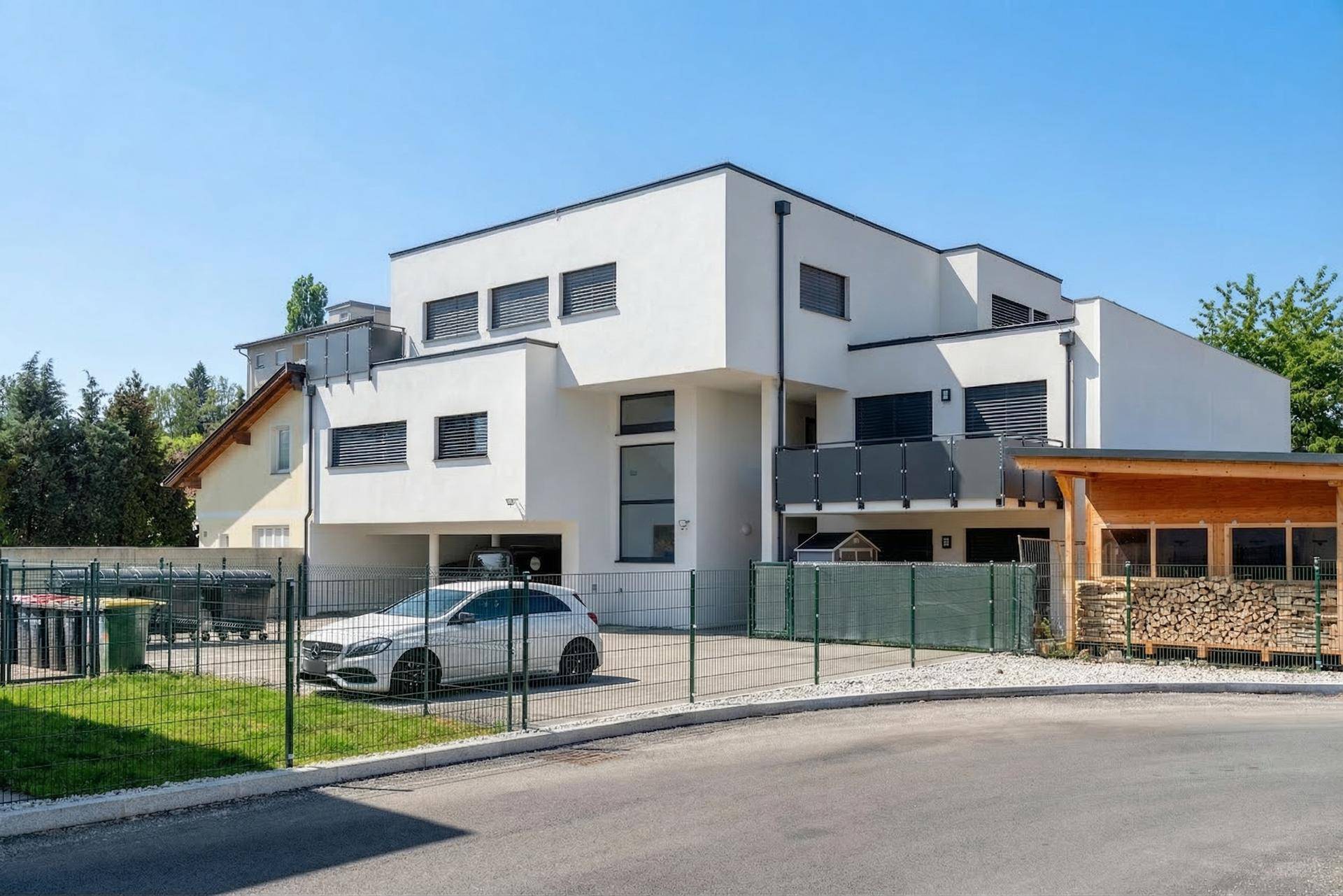 Modernes Mehrfamilienhaus mit Parkplätzen, Carport und Holzstapel im Außenbereich unter blauem Himmel.