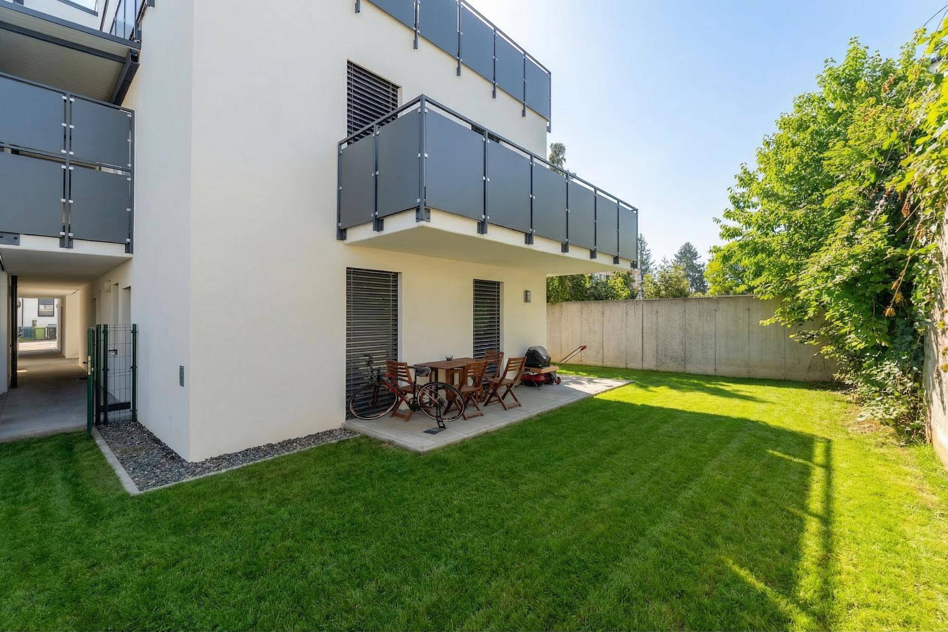 Sonniger Gartenbereich mit gepflegtem Rasen und einer gepflasterten Terrasse mit Gartenmöbeln.
