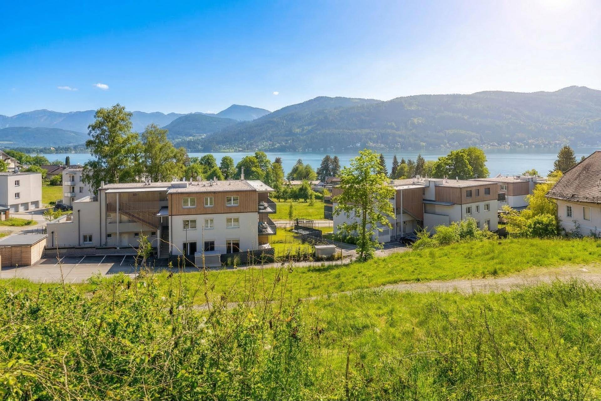 Panoramablick von einer grünen Wiese auf moderne Gebäude, einen See und die umliegende Berglandschaft.