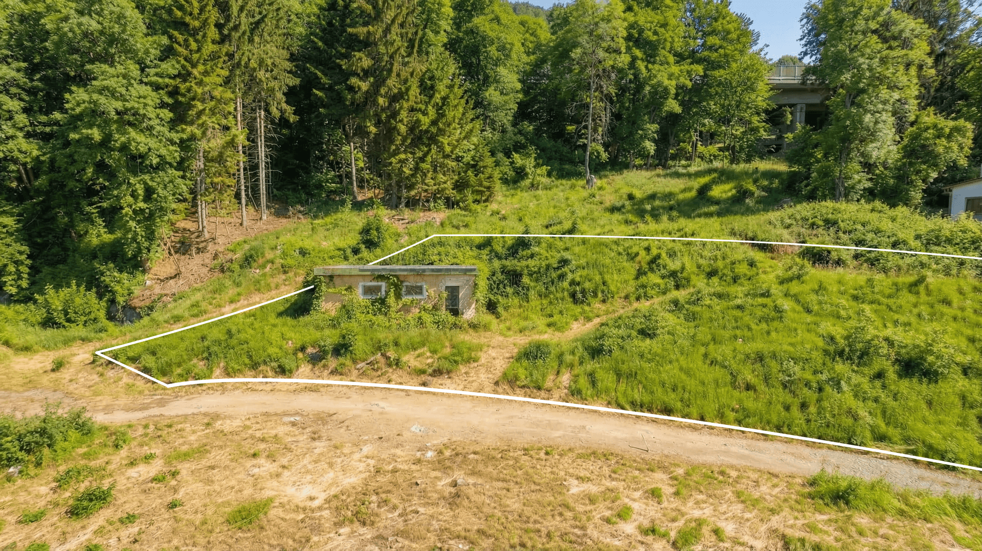 Blick auf einen grasbewachsenen Hang mit einem kleinen Gebäude, einem Feldweg und einem markierten Grundstück.