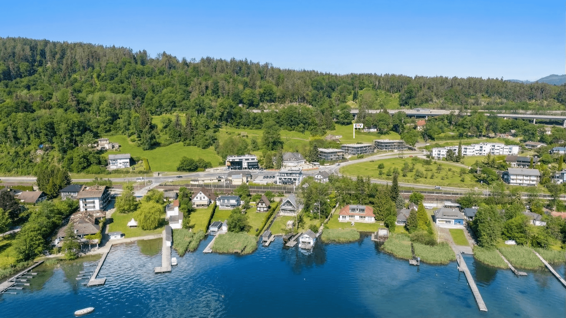Panoramablick auf eine Stadt am Seeufer mit Häusern, Stegen und einer Straße vor Bergkulisse.