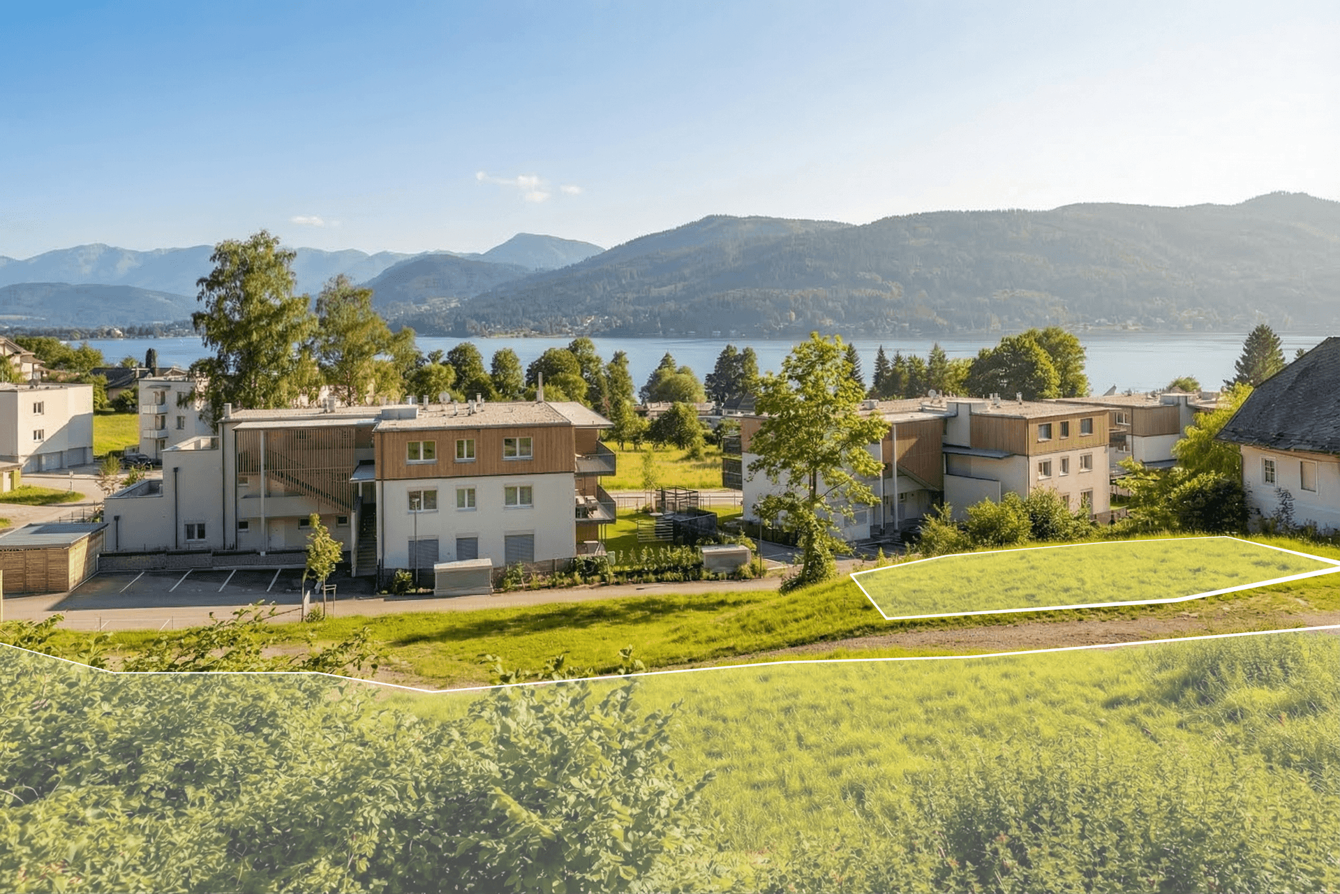 Blick von einer Wiese auf moderne Gebäude, einen See und Berge, mit einem markierten Grundstück im Vordergrund.
