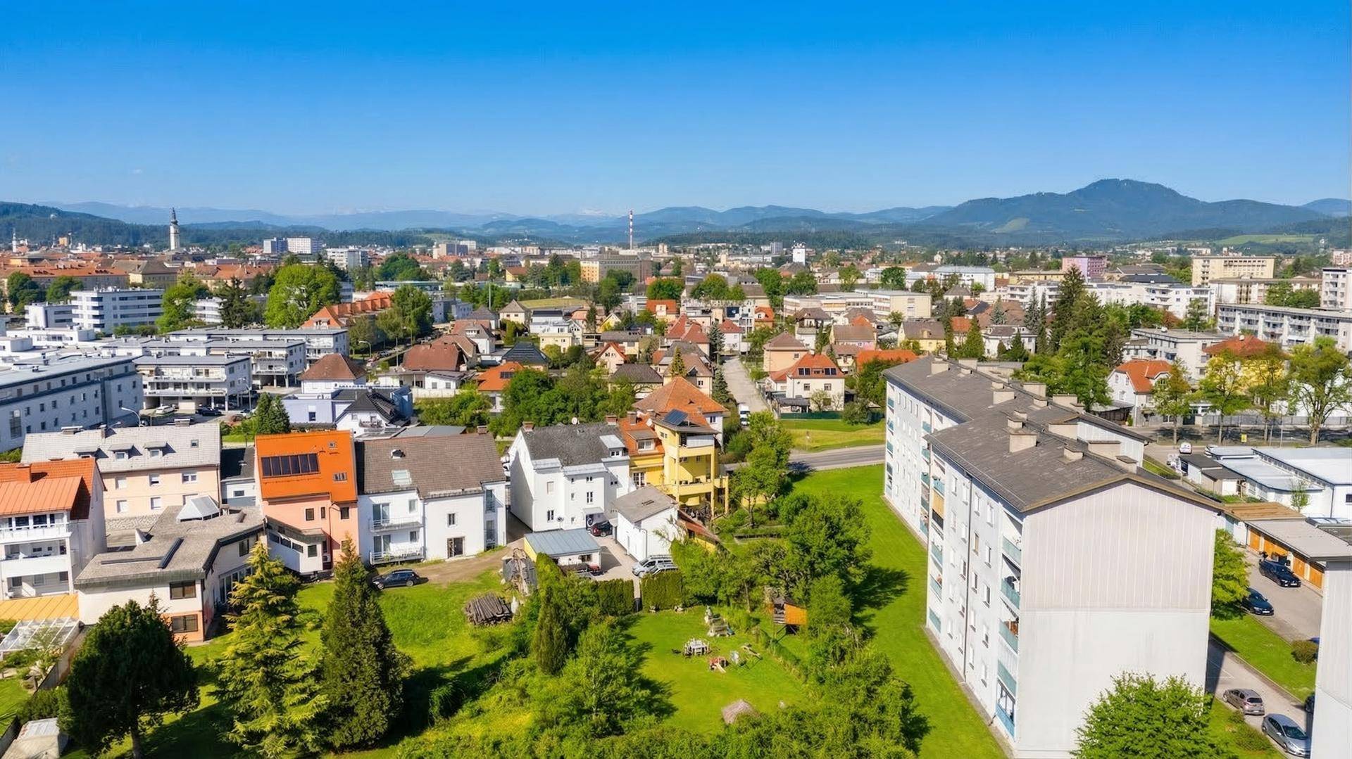 Weitläufige Vogelperspektive einer Stadt mit Wohngebäuden, Grünflächen und Bergen am Horizont.