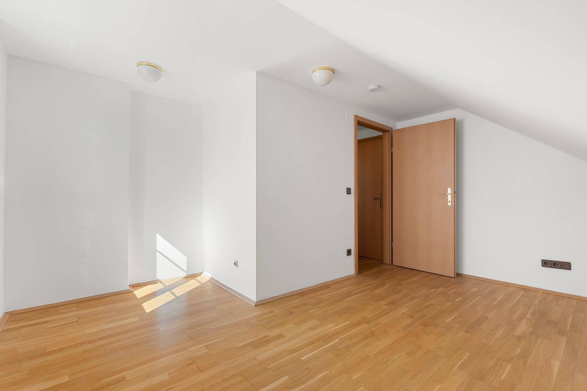 Helles Zimmer mit Holzparkettboden, weißen Wänden und Dachschrägen, bereit zur individuellen Gestaltung.