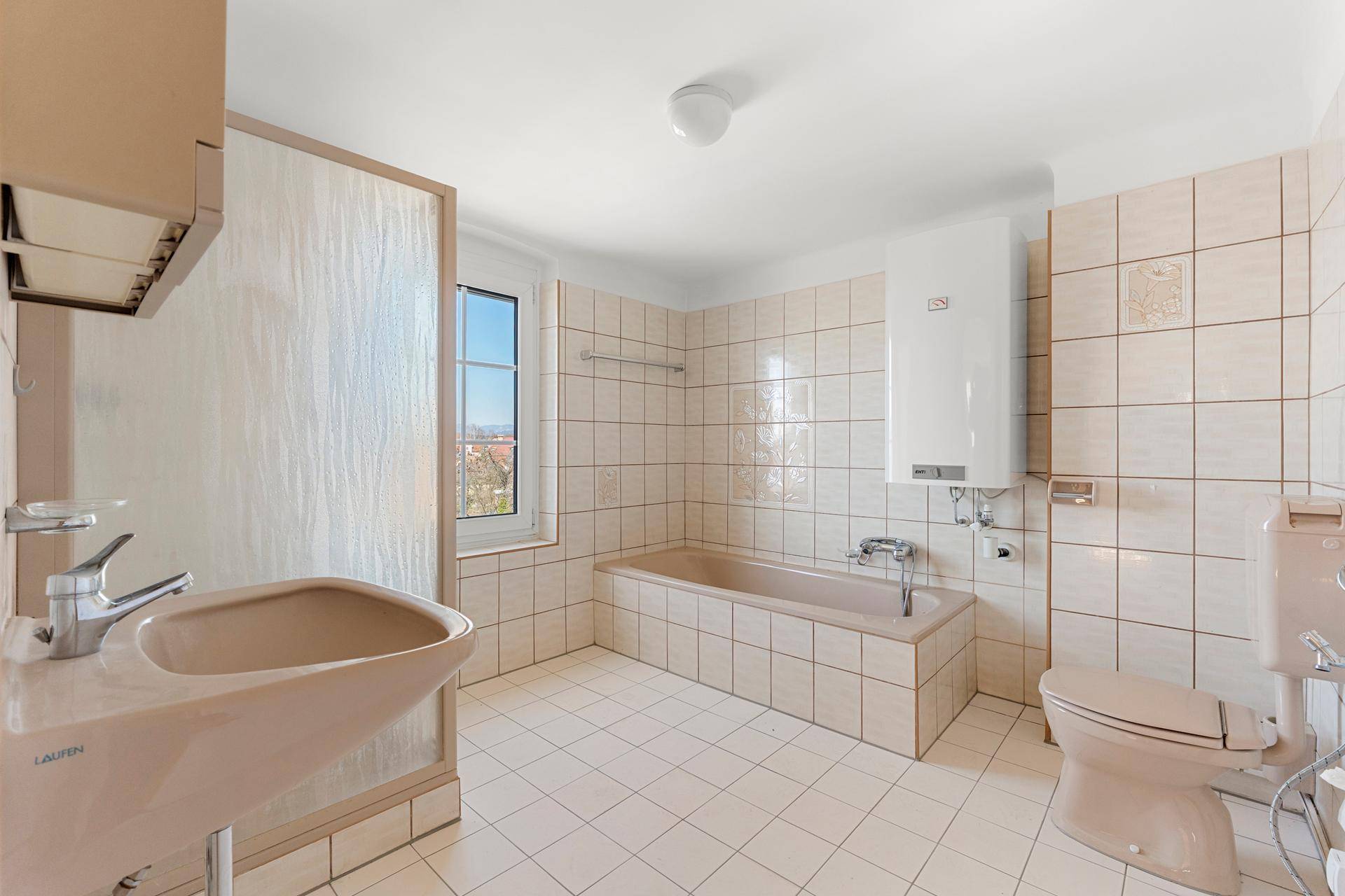 Badezimmer mit beigefarbenen Fliesen, Badewanne, Waschbecken und Fenster, im Stil der 80er Jahre.